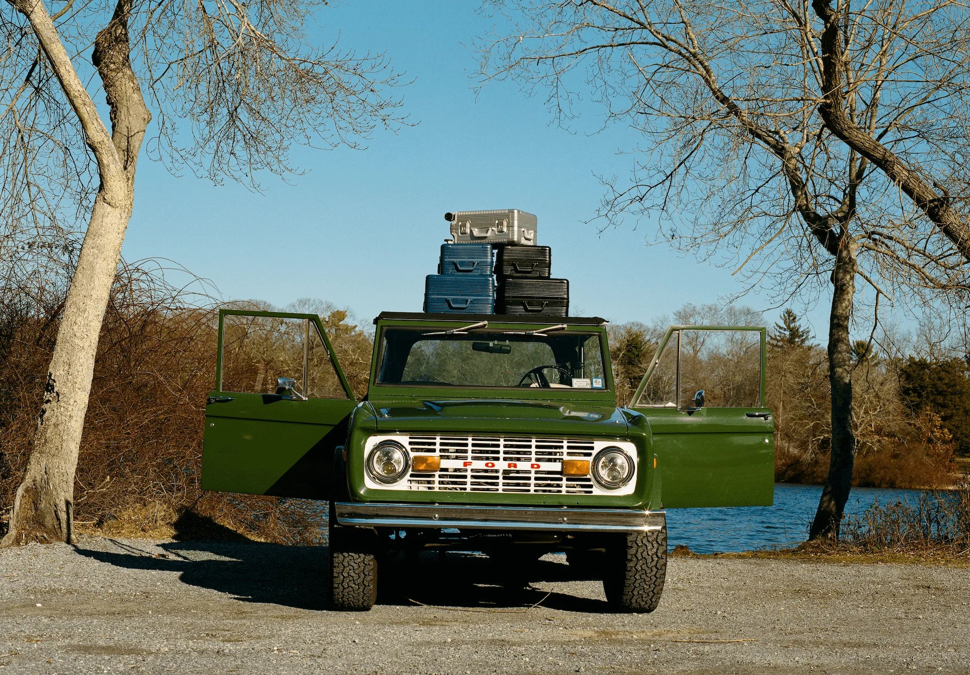 A vintage green truck parked by a lake, with luggage stacked on top and trees in the background.